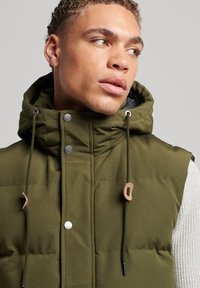 Wattierte grüne Weste mit Kapuze, mit Druckknöpfen, Schnürung und einem strukturierten weißen Pullover darunter.