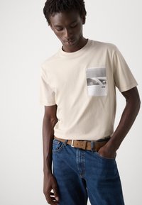Beige bomulls t-skjorte med grafisk lomme detalj, kombinert med blå denim jeans og et brunt belte. T-skjorten har rund hals og korte ermer.