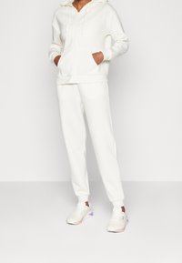 Felpa bianca con tasche frontali e cordini, abbinata a pantaloni della tuta bianchi. Le scarpe sono sneakers chiare con accenti pastello.