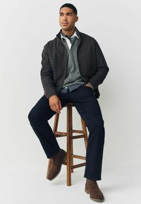 Pánská hnědá bunda na zip přes šedý svetr, doplněná tmavými kalhotami a hnědými semišovými botami. Sedí na dřevěné stoličce na světlejším pozadí.