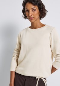 Sweatshirt couleur crème à col rond et manches longues, avec une texture lisse et une coupe décontractée. Associé à un pantalon marron foncé.