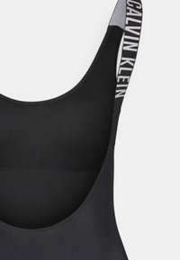 Maillot de bain noir avec un décolleté en cœur et des bretelles larges. Les bretelles présentent un motif de logo contrastant. Le matériau semble lisse et élastique.