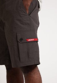 Shorts gris avec une texture froissée, dotés d'une poche cargo sur le côté et d'une étiquette rouge "JACKIT". Ils ont un détail de fermeture à bouton.