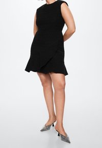 Schwarzes ärmelloses Kleid aus strukturiertem Stoff, mit asymmetrischem Vorderteil und gerafftem Saum. Kombiniert mit silberakzentuierten spitzen Absatzschuhen.