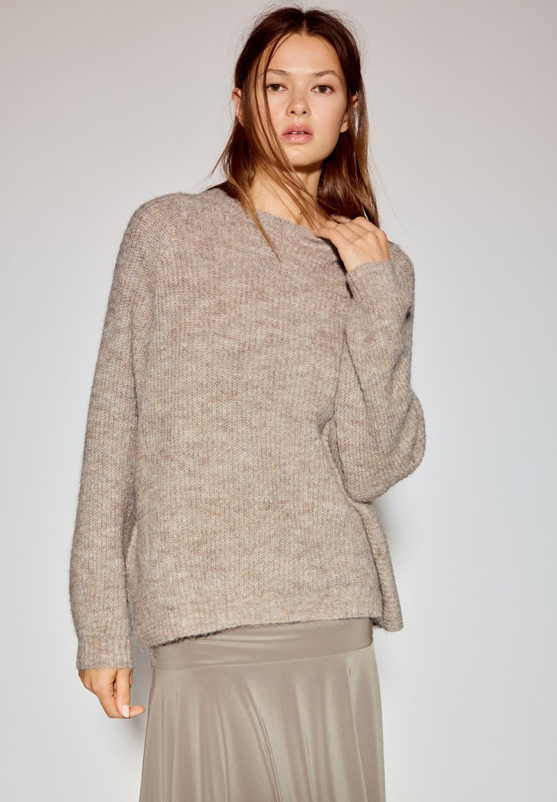 Béžový pletený sveter s voľným strihom, okrúhlym výstrihom a rebrovanými manžetami, vrstvený cez hodvábnu, svetlohnedú sukňu. Jemná textúra a ležérny dizajn.