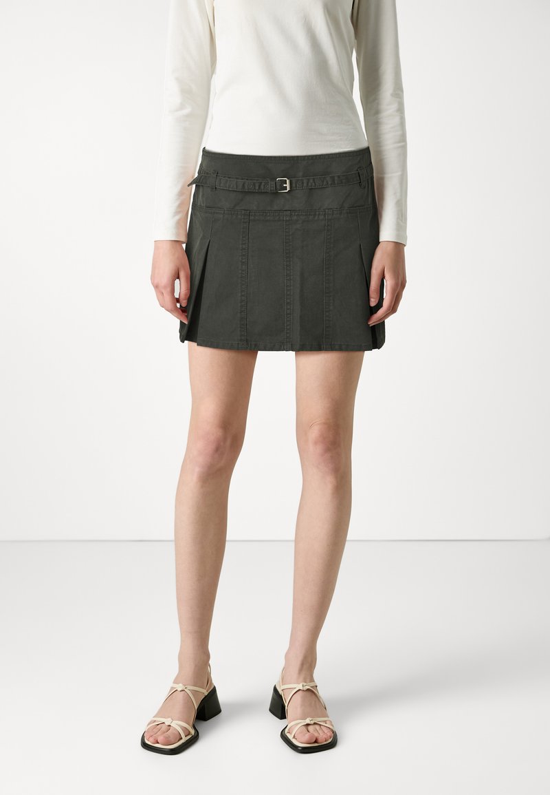 Jupe en denim gris foncé avec une taille ceinturée, un design plissé et des détails de couture verticaux. Assortie avec des sandales beiges à lanières.
