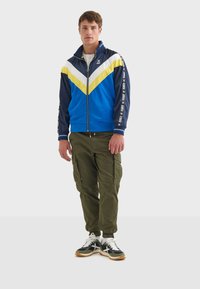 Blå och gul zip-up träningsjacka med chevrondesign, tillsammans med olivgröna cargobyxor och svarta sneakers med vita accenter.