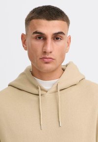 A young man with short hair wears a beige hoodie over a white shirt, staring directly at the camera with a neutral expression.