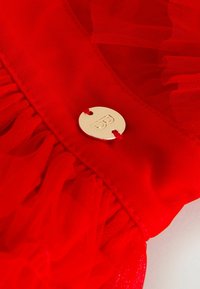 Červená tkanina z tulového materiálu s volánovými okrajmi; obsahuje kruhový zlatý kovový doplnok s nápisom „B.“ Výrazná textúra a žiarivá farba.