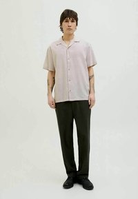 Jovem com cabelo curto e escuro, usando uma camisa de botão de manga curta, bege claro, calças escuras e sapatos pretos, de pé contra um fundo simples.