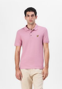 Lys rosa polo skjorte med klassisk krage, tre knapper foran og korte ermer. Har en liten gul sommerfugl-logo på brystet.