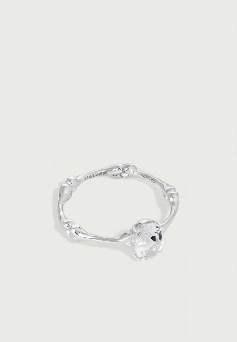 Bague en argent avec un anneau sculptural et irrégulier, ornée d’une grande pierre précieuse ovale et transparente, sur un fond clair uni.
