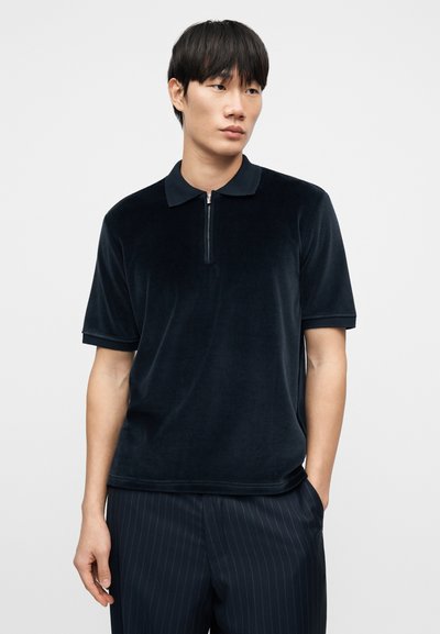 Μπλε ναυτικό βελούδινο πόλο πουκάμισο με φερμουάρ στον λαιμό, κοντά μανίκια και γιακά. Το ύφασμα φαίνεται απαλό και λείο, ενισχύοντας τη συνολική άνεση.