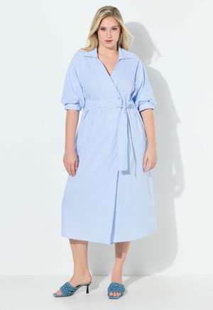 Kobieta w jasnoniebiesko-białej paskowanej sukience kopertowej z rękawami 3/4 i wiązanym w pasie, w zestawieniu z niebieskimi plecionymi sandałami na wysokim obcasie.