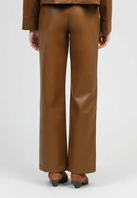Pantalons larges en simili cuir marron avec une texture lisse, présentant un ourlet net et un design sans embellissement. Portés avec des chaussures à talons marron.