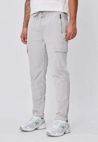 Homme portant un pantalon cargo gris clair avec taille à cordon et poche zippée, associé à des baskets blanches New Balance, debout devant un fond uni.