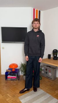 Μαύρη μπλούζα Nike με κόκκινο λογότυπο, φαρδιά σκούρα τζιν παντελόνια, γυαλιστερές μπορντό loafers. Στηρίζει σε ξύλινο πάτωμα με πολύχρωμη διακόσμηση και τηλεόραση.