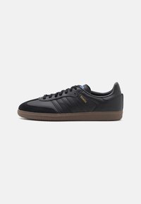 Black leather sneakers featuring three tonal stripes, a textured black toe cap, and a brown rubber sole. "SAMBA" logo displayed in gold.