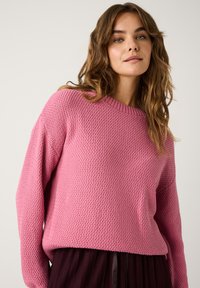 Texturierter rosa Pullover mit Rundhalsausschnitt und fallenden Schultern, kombiniert mit einem dunklen, gefalteten Rock. Klarer, leichter Strickstoff.