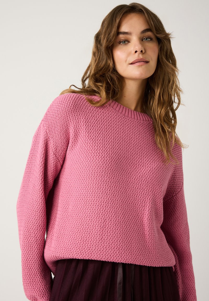 Texturierter rosa Pullover mit Rundhalsausschnitt und fallenden Schultern, kombiniert mit einem dunklen, gefalteten Rock. Klarer, leichter Strickstoff.