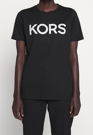 Personne portant un T-shirt noir à manches courtes avec de grandes lettres blanches cloutées formant le mot « KORS » sur la poitrine.