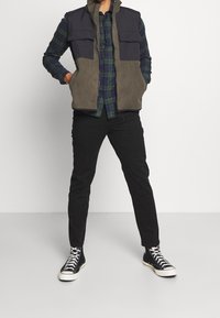 Mans väst i marinblått och olivgrönt med två framfickor, bärs över en grön och marinblå rutig skjorta, tillsammans med svarta byxor och hög sneakers.