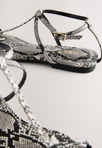 Platte sandalen met zwart-wit slangenhuidpatroon, dunne bandjes en een gouden gesp op een neutrale achtergrond.