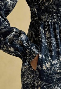 Námornícko modré šaty s bielym kvetinovým vzorom, riasený trup a elastické manžety. Obsahuje bočnú vrecko na boku. Mäkká textúra látky.