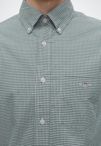 Chemise à carreaux verts et blancs avec boutons et une poche avant ornée d'un petit logo écusson brodé.