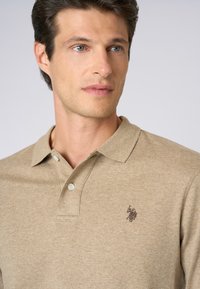 Uomo con occhi azzurri chiari che indossa una polo beige con bottoni e un piccolo emblema scuro ricamato sul petto, che guarda leggermente a destra.