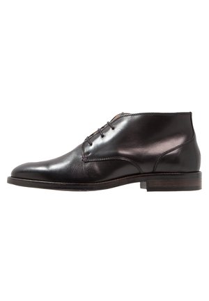 Zapato de vestir para hombre de cuero negro para el tobillo con cordones, punta ligeramente puntiaguda y tacón bajo apilado sobre fondo blanco.