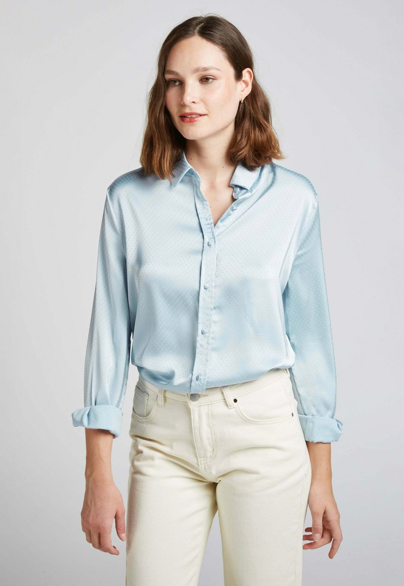 Chemisier satiné bleu clair avec un devant boutonné, des manches longues retroussées aux poignets et un motif en petits pois subtil sur l'ensemble. Associé à un pantalon clair.