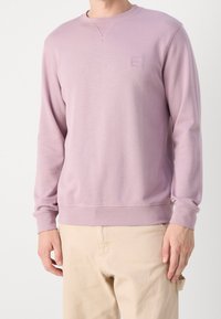 Fialová mikina s kulatým výstřihem a žebrovanými manžetami. Na hrudi má malou našitou značku. Měkká látka s hladkou texturou.