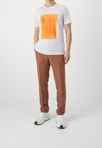 Weißes Leinen-T-Shirt mit einem orangen rechteckigen Grafik, kombiniert mit braunen Leinenhosen und weißen Sneakers mit schwarzen Akzenten.