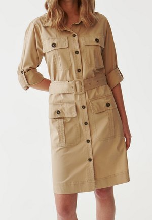 Robe-chemise beige boutonnée, manches retroussées, deux poches poitrine, deux grandes poches basses et une ceinture assortie à la taille.