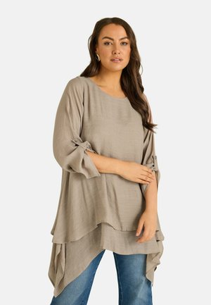 Donna con tunica beige larga a strati con maniche arrotolate e jeans blu, in piedi davanti a uno sfondo chiaro e semplice.