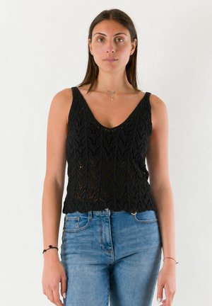 Donna con capelli castani lisci indossa un top nero senza maniche lavorato a maglia e jeans blu, in piedi davanti a uno sfondo chiaro uniforme.