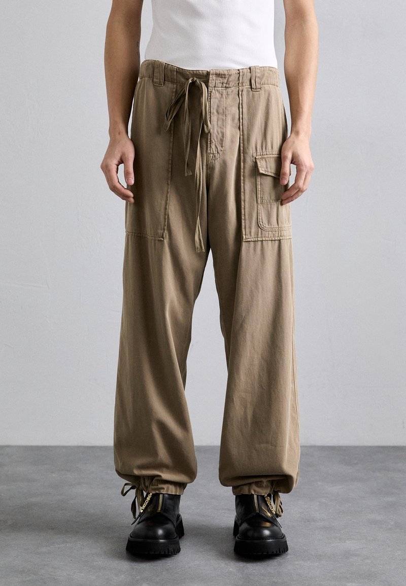 Personne portant un pantalon cargo beige ample avec une taille et des bas à cordon de serrage, assorti de bottes noires, debout sur un sol gris.