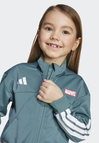 Teal track jacket with white stripes, zip front, and red "MARVEL" logo on the chest. Made of smooth, lightweight material.
