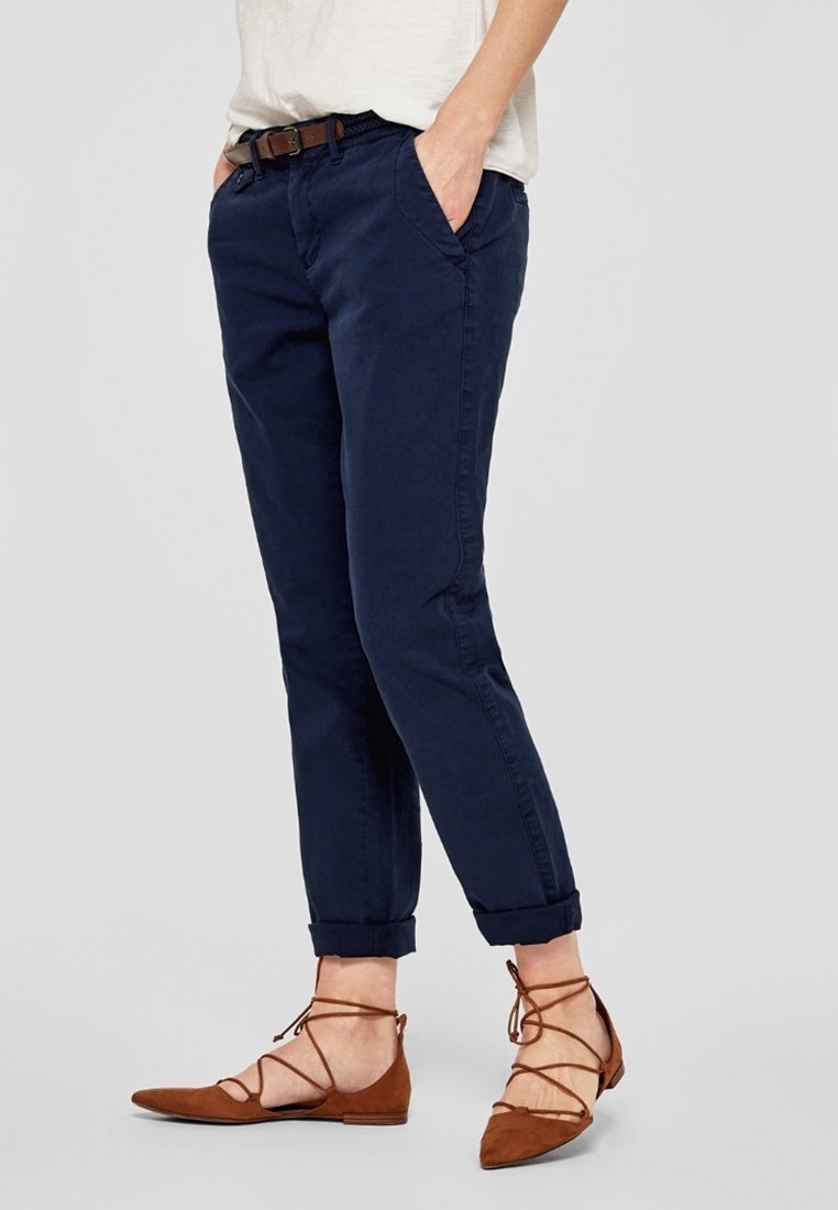 Personne portant un pantalon bleu foncé roulé, un haut blanc, une ceinture marron et des chaussures plates marron à lacets, les mains dans les poches.