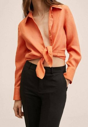 Vrouw draagt een oranje blouse met lange mouwen, gestrikt in de taille, en zwarte broek met hoge taille, met één hand in de zak.