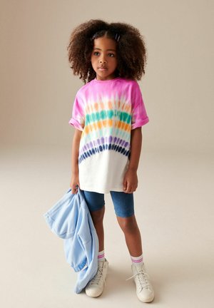 Kortærmet tie-dye t-shirt i pink, hvid, grøn, orange og lilla, parret med denimshorts og hvide sneakers, mens der holdes en blå sweatshirt.
