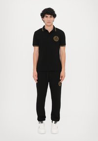 Polo negru cu accente galbene, având un logo brodat; asociat cu pantaloni de trening negri. Modelul poartă adidași albi.