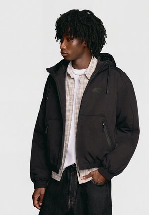 Junger Mann mit dunklem lockigem Haar, der einen schwarzen Kapuzenpullover mit Reißverschluss über einem beige karierten Hemd und weißem T-Shirt trägt und mit einer Hand in der Tasche steht.