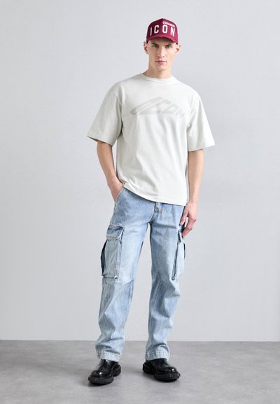 T-shirt surdimensionné blanc avec logo gris, pantalon cargo bleu clair avec poches latérales, chaussures noires épaisses et casquette bordeaux avec lettrage blanc.