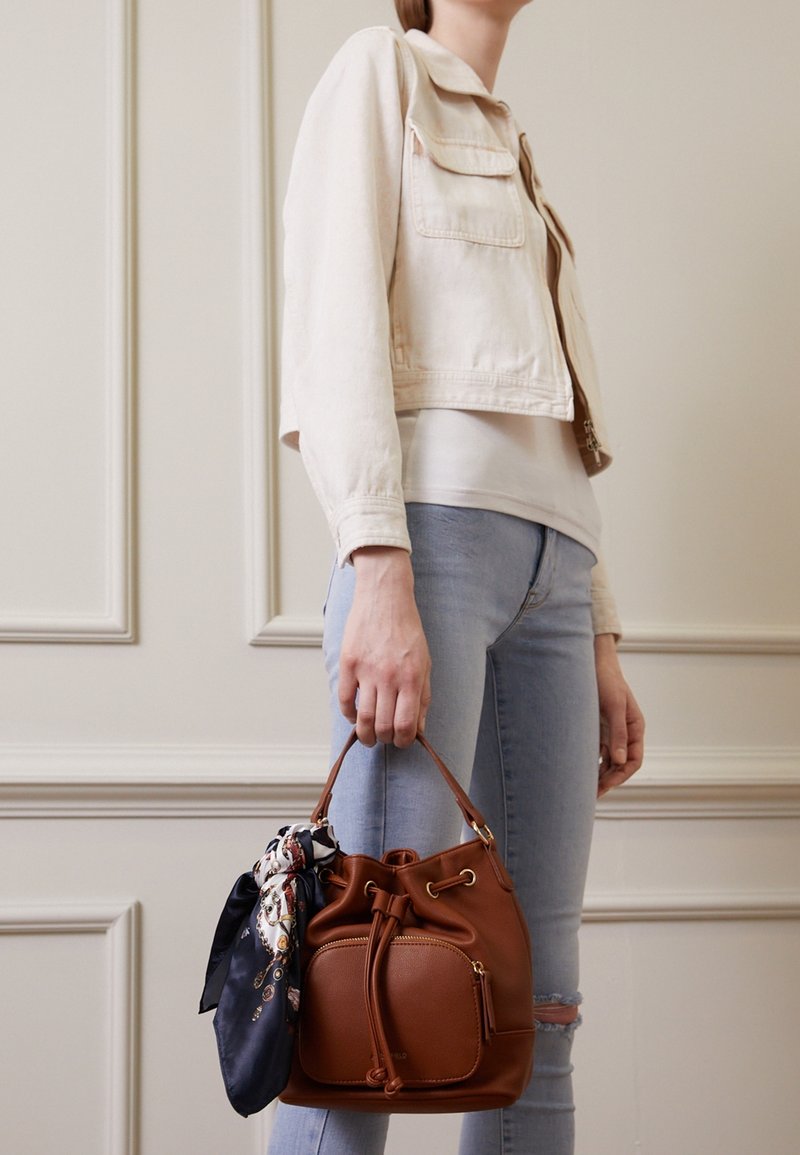 Femme portant une veste légère et un jean, tenant un sac seau en cuir marron avec un foulard à motifs attaché à la poignée.