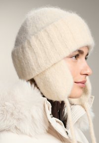 Krémová pletená čiapka s klapkami na uši, vyrobená z jemnej vlny. Má rebrovanú textúru a šnúrky, doplnená nadýchaným bielym kabátom s kožušinovou ozdobou.