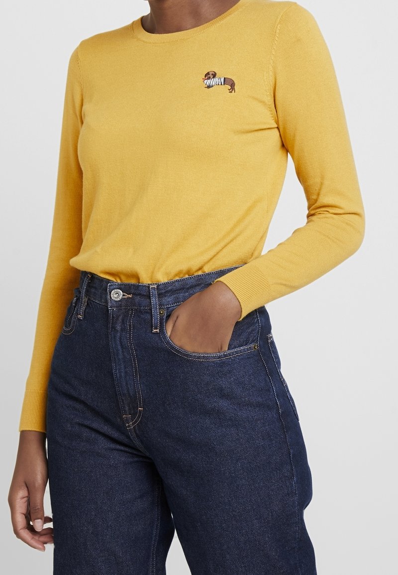 Personne portant un pull jaune à manches longues avec un petit chien brodé et un jean taille haute bleu foncé, main dans la poche.