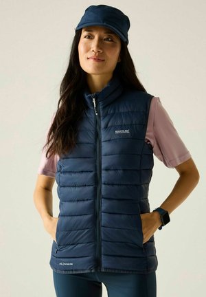 Vrouw die een marineblauwe gewatteerde bodywarmer en een pet draagt, lichtpaars T-shirt met korte mouwen en een smartwatch, staand met handen in de zakken van de bodywarmer.