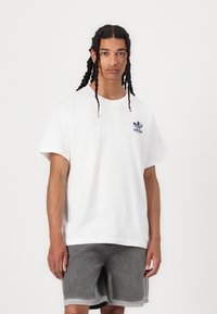 Hvit bomulls t-skjorte med blå Adidas-logo på brystet, rund hals og korte ermer. Kombinert med grå denimshorts.